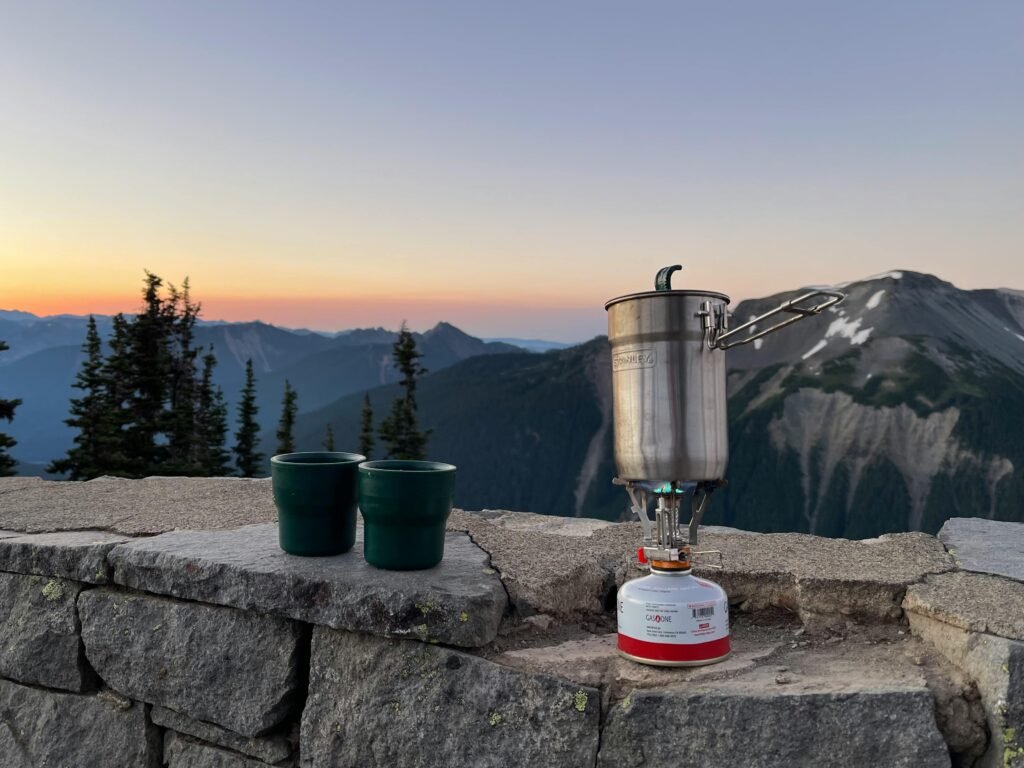 A backpacker using a compact jet boil stove to quickly boil water for a meal against a scenic mountain backdrop.