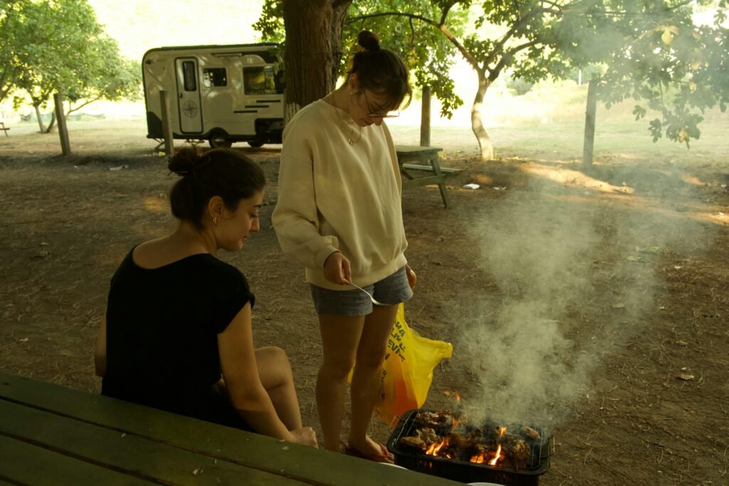 A person grilling food on a portable Coleman BBQ stove during a sunny day at a campsite, showcasing the convenience of outdoor cooking.