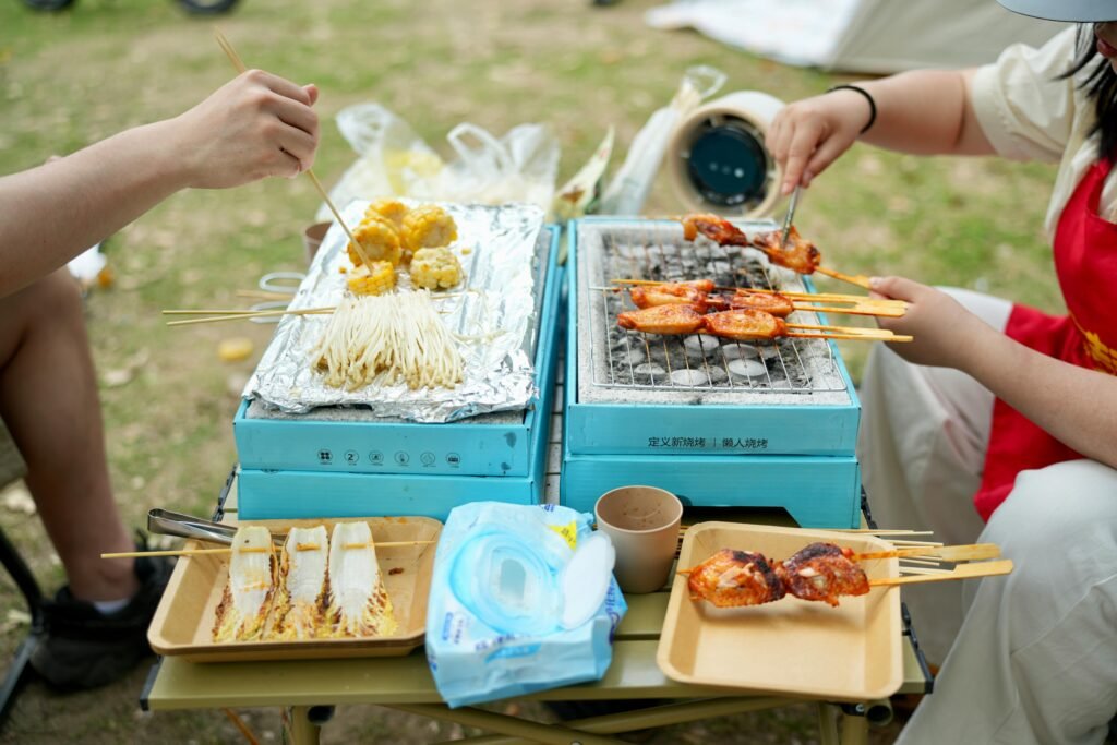 A person grilling burgers and vegetables on a red Coleman portable grill at a sunny campsite in the woods.