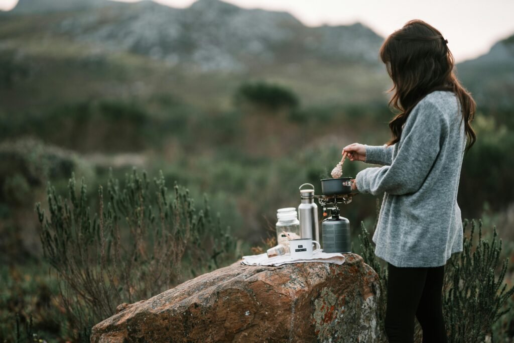 A person cooking a meal on a modern portable top stove at a campsite with a scenic mountain and forest landscape in the background.