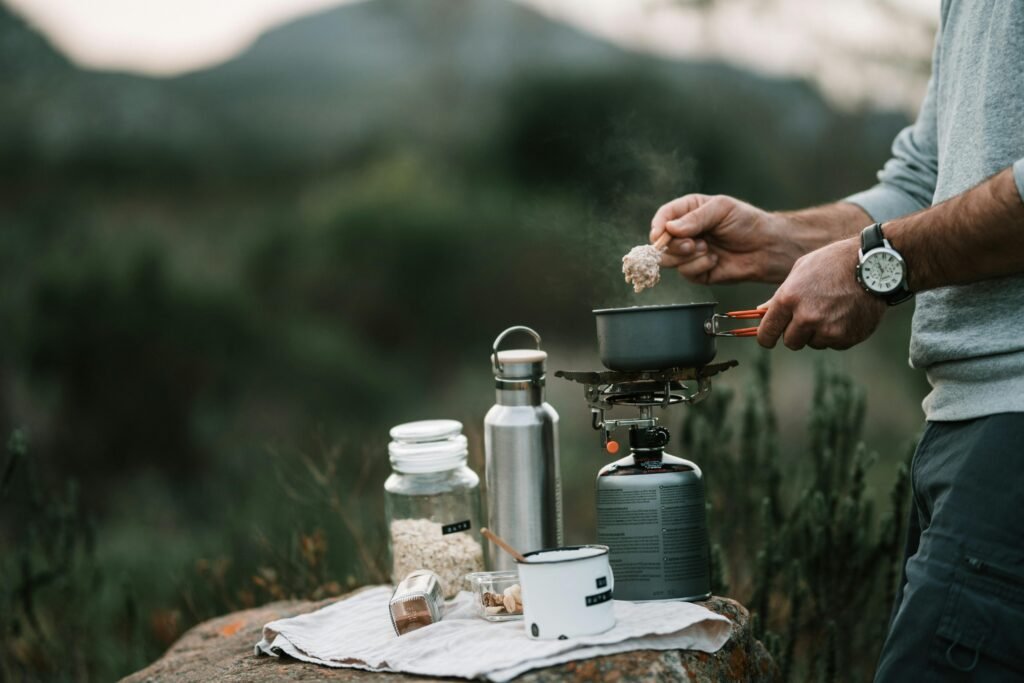 A person cooking a meal on a modern two-burner outdoor propane stove set up on a picnic table at a scenic campsite.
