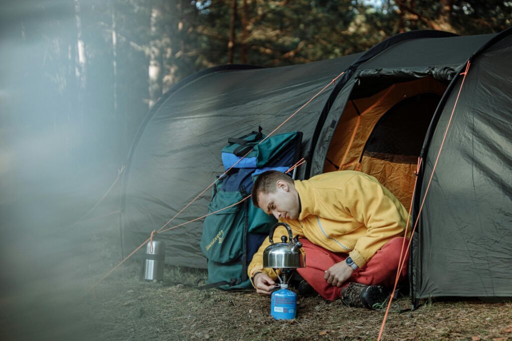 A person cooking breakfast on a classic two-burner propane camping stove at a campsite surrounded by trees.
