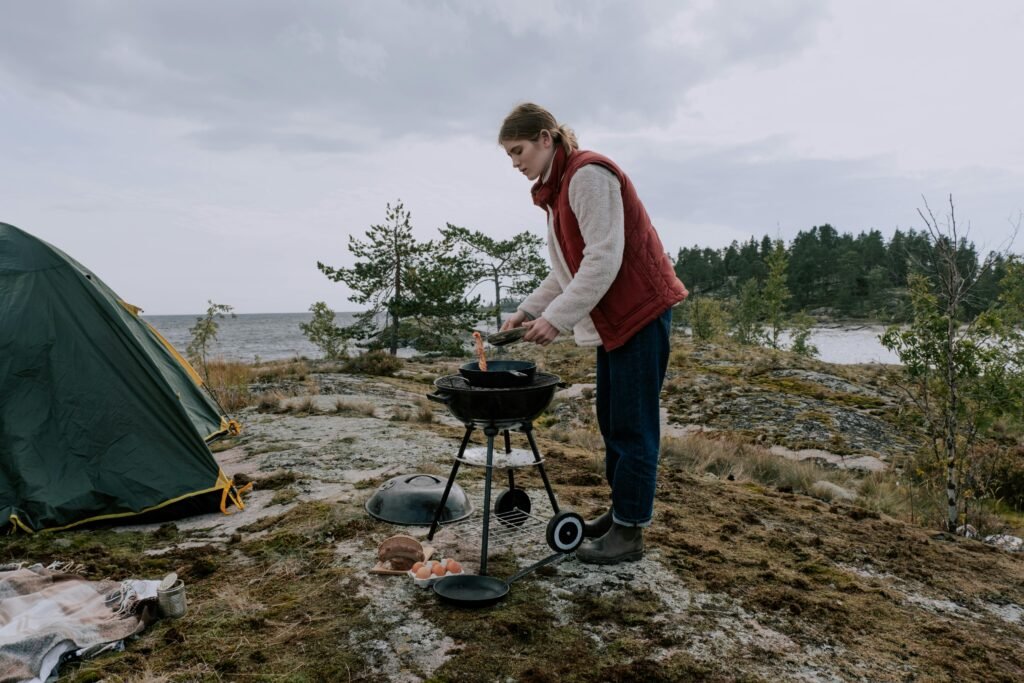 A camper uses a compact dual fuel camp stove to prepare a hot meal in a scenic mountain setting, highlighting its versatility for outdoor cooking with white gas or unleaded gasoline.