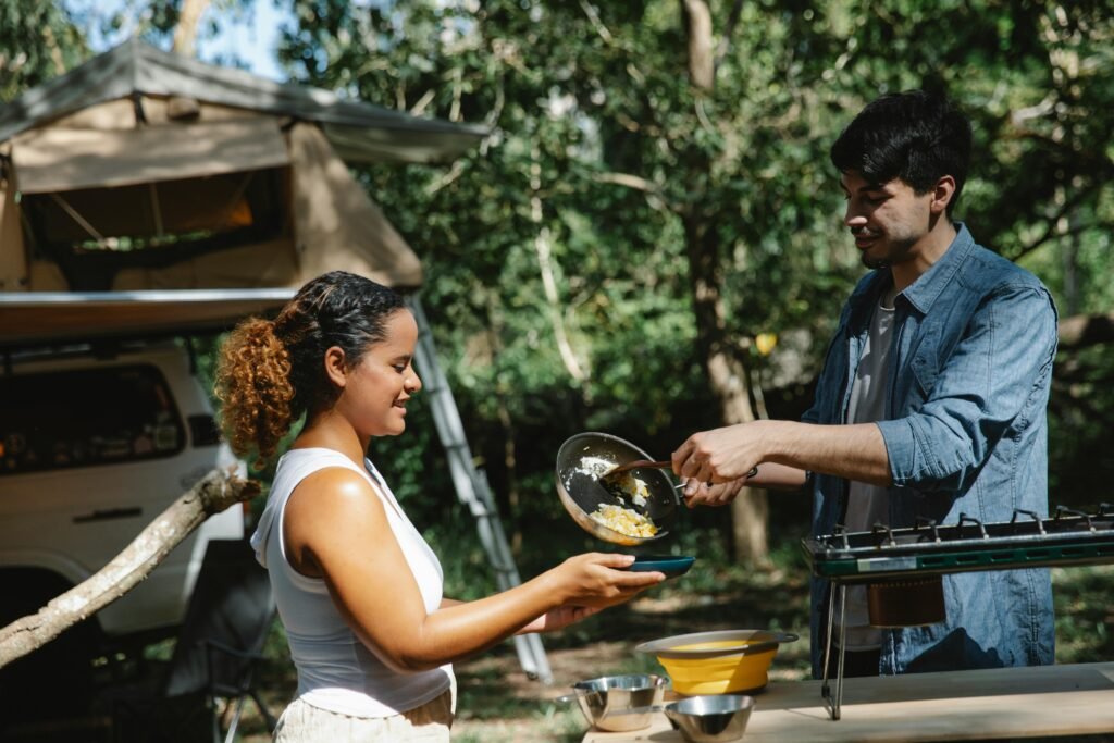 A person cooking breakfast on a classic green Coleman propane stove at a beautiful campsite in the woods.