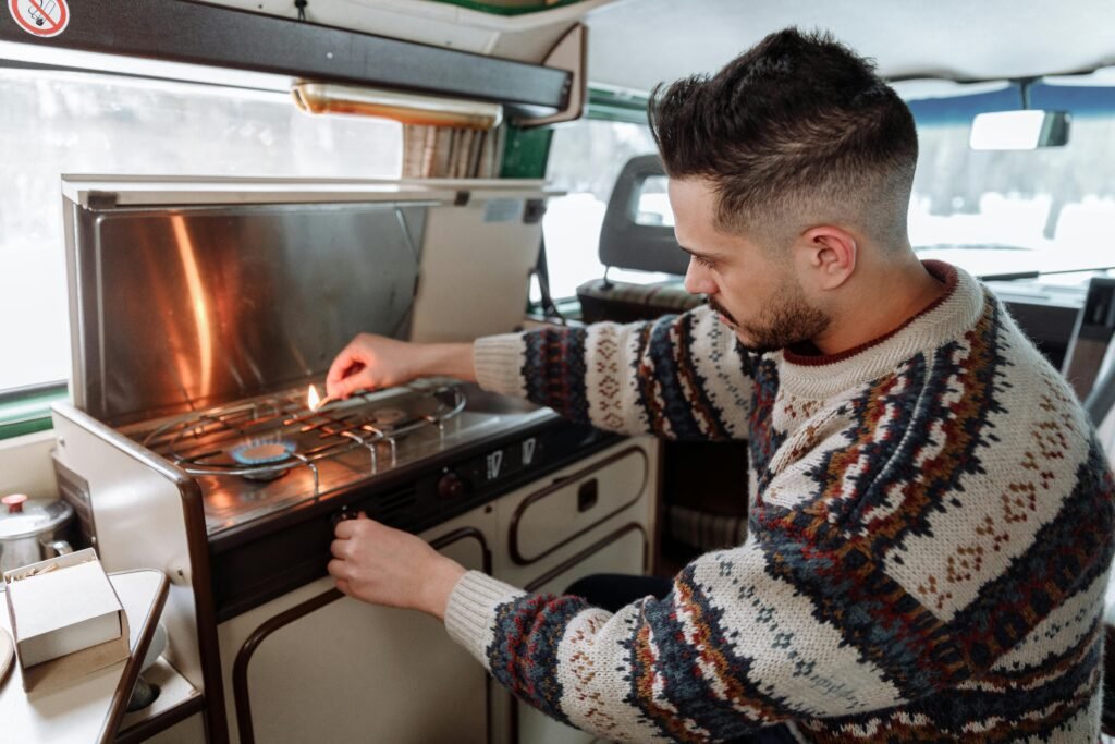 A person cooking breakfast on a portable camper gas stove, set up on a wooden table at a scenic campsite.