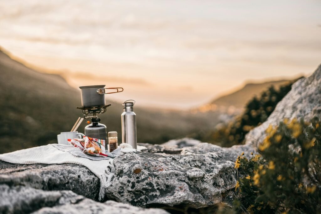 A person cooking a meal on a small, portable propane stove at a campsite with a scenic mountain view in the background.